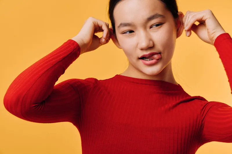 Young woman with dark hair wearing red sweater adjusting her hair against orange background. Casual style, thoughtful expression, portrait for fashion or lifestyle concept.