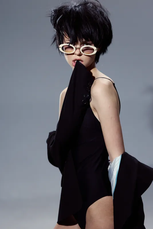 Stylish woman with short black hair in black outfit and white sunglasses posing against gray background.