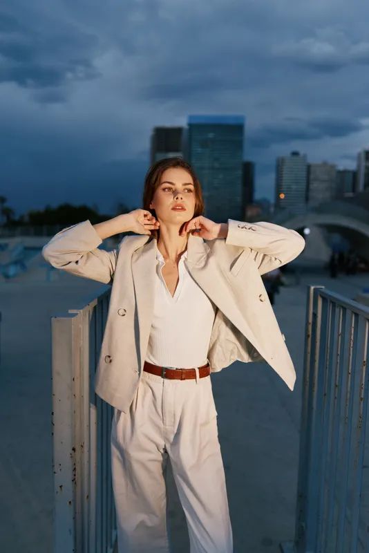 Confident woman in beige business suit posing outdoors at dusk with modern city skyline background, showcasing fashion, style, and empowerment