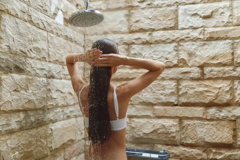 Woman showering under rain showerhead in stone bathroom, long wet hair, relaxed posture, natural light, minimalistic spa atmosphere, warm earthy colors.