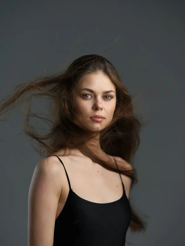Portrait of a young woman with flowing brown hair wearing a black dress against a gray background
