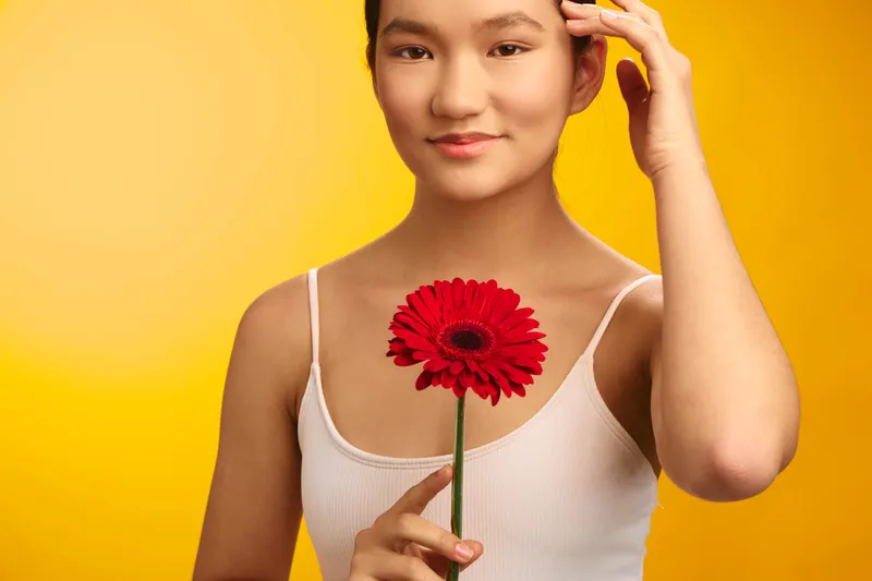 asian woman holds a red flower against a vibrant yellow background, presenting a warm, calm portrait with a friendly gaze and graceful, natural poise.