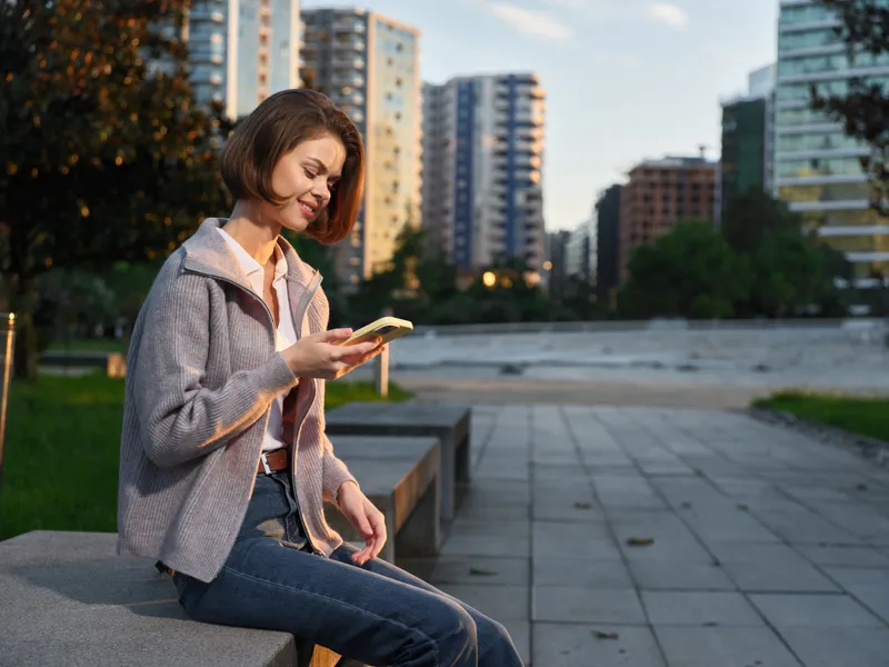 A cheerful woman sits on a park bench outdoors, texting on her phone with a friendly smile, enjoying a sunny day in an urban setting, surrounded by modern buildings and a light breeze.