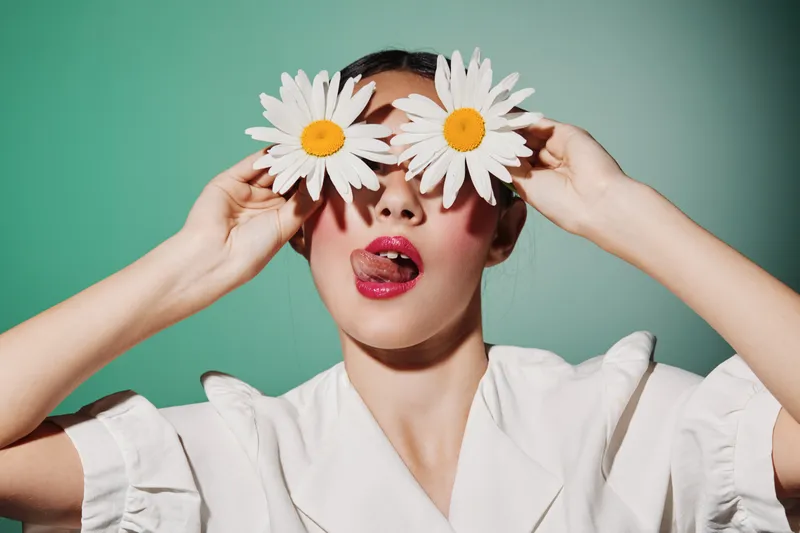 Young woman holds two daisies over her eyes playfully, sticking out her tongue. She wears red lipstick and a white blouse against a clear pastel backdrop. Concept of beauty fun and fashion.