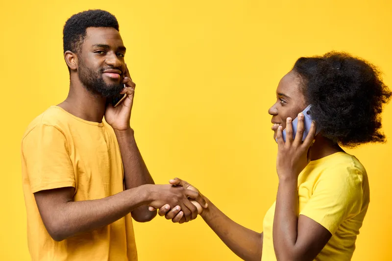 Young African couple communicating over the phone, smiling and shaking hands against a bright yellow background, showcasing positivity and connection in modern relationships