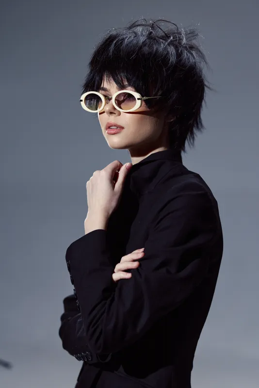 Stylish woman with short black hair wearing sunglasses and a black turtleneck expresses confident elegance.
