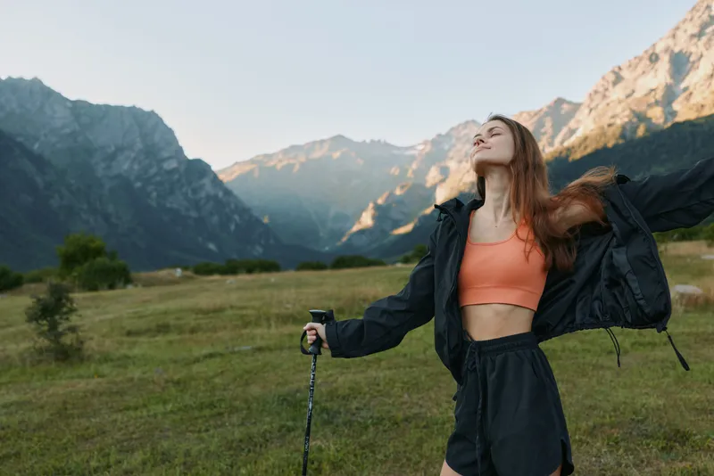 A young woman stands in a grassy valley with mountains in the background, wearing a coral crop top and dark jacket, arms outstretched, embracing freedom and adventure in nature.