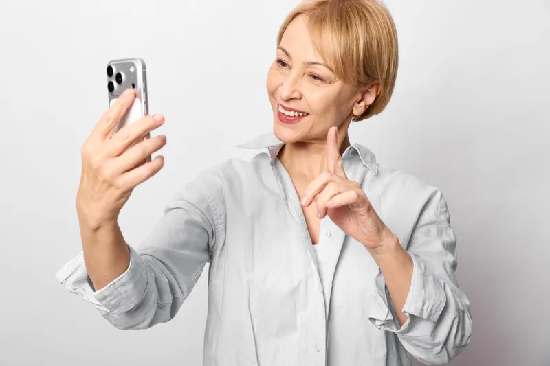 Elderly woman holds smartphone for a selfie, smiling in portrait view, communicating through technology for social lifestyle, indoors on white clean background