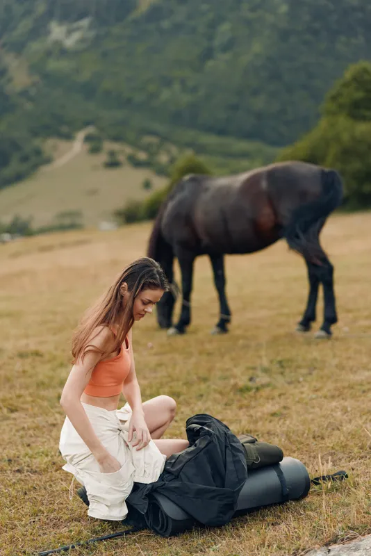 A young woman prepares gear in a grassy meadow while a horse grazes nearby. The scene captures outdoor leisure, nature, and a calm rural atmosphere.
