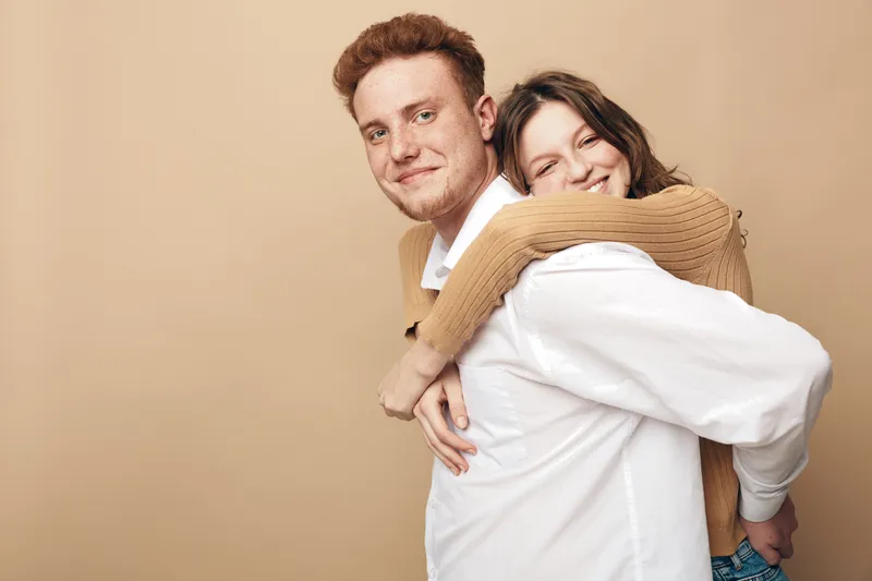 Happy young couple with woman hugging man from behind, both smiling and looking at camera. Casual clothing and warm beige background create cozy romantic mood.