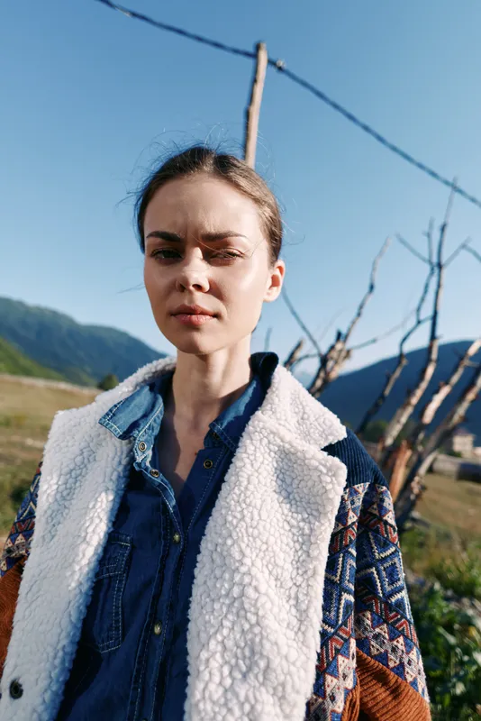 Woman portrait outdoors in mountains and rural field wearing a denim shirt and warm jacket under sunlight, serious gaze and natural expression in scenic countryside landscape.