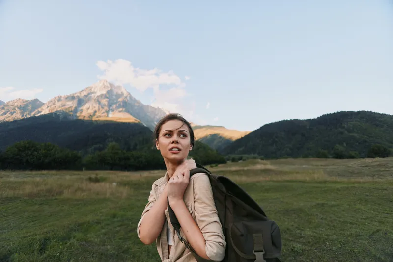 Woman with backpack standing in an open field near mountains, embracing outdoor life, looking ahead with a determined expression, capturing daily adventure and countryside serenity.