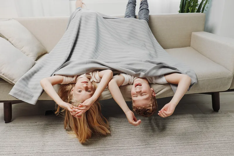 Two happy children lying upside down on a sofa covered with a grey blanket in a bright living room. Concept of family fun, relaxation, and cozy home moments together.