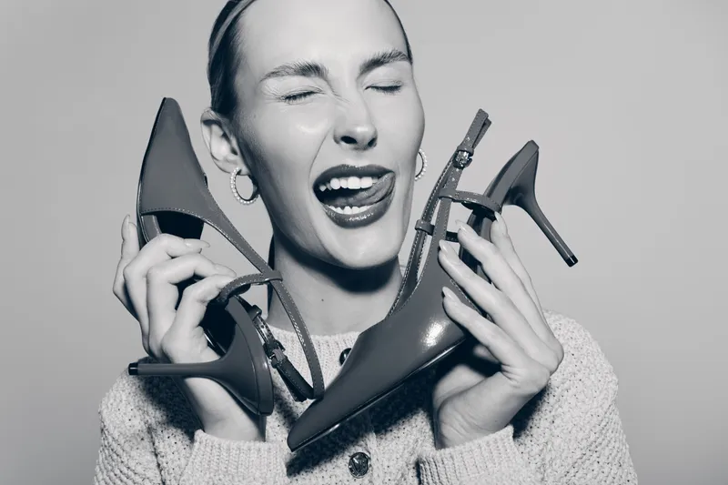 Young Caucasian woman with light skin holds high heel shoes near face, smiling with closed eyes and playful expression.