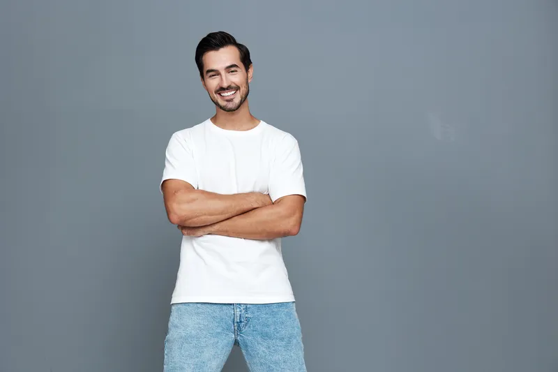 Smiling young man standing against gray background with arms crossed, wearing casual white t-shirt and blue jeans, exuding a cheerful and relaxed atmosphere.