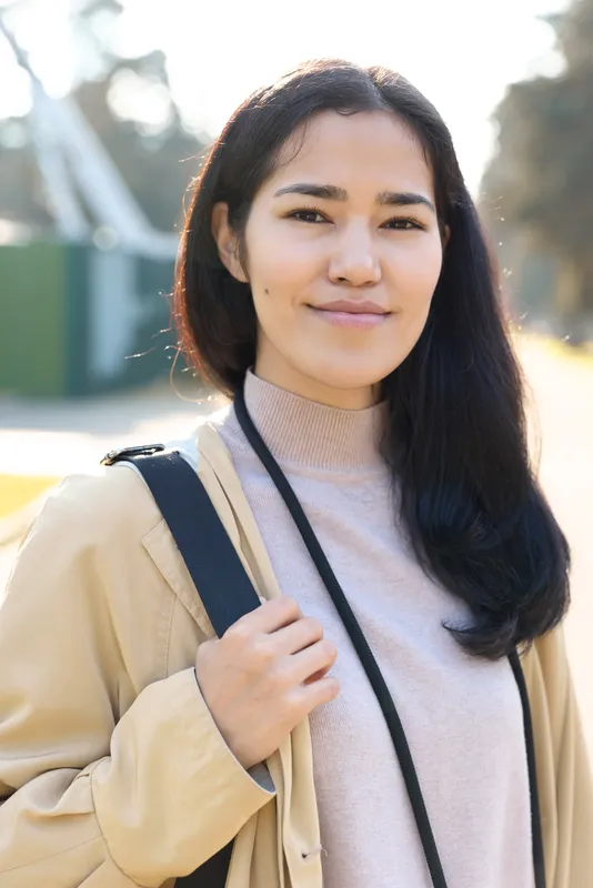 Smiling woman outdoors in a beige coat and soft sweater, carrying a shoulder bag, sunlit backdrop, casual fashion and relaxed mood for everyday lifestyle and travel moments