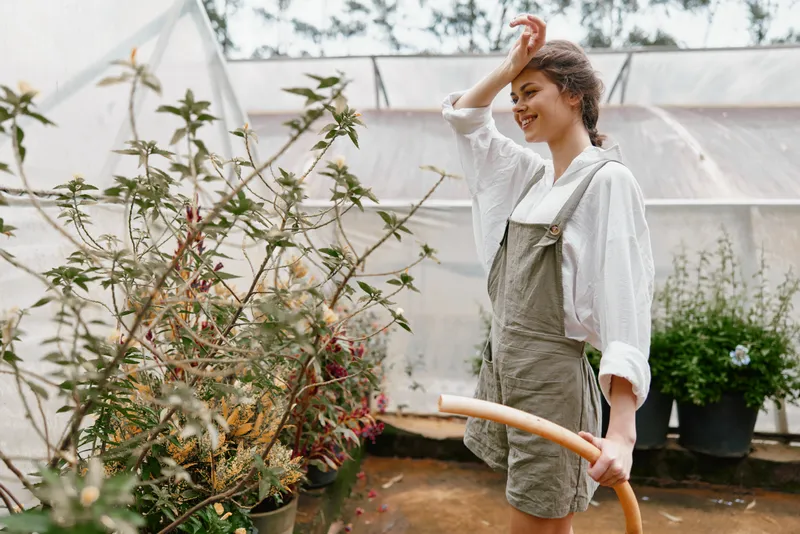 Young woman gardening in a greenhouse, wearing a minimalist outfit and smiling while watering plants, showcasing a connection to nature and sustainable living