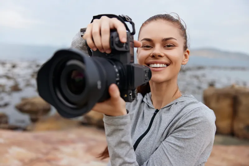 Cheerful young woman smiling and holding a camera outdoors, showcasing her photography skills against a scenic natural backdrop with soft colors