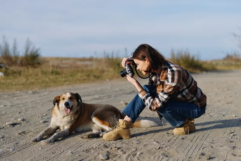 Photographer, kneeling woman with camera, captures a candid outdoor moment with a dog on a sandy trail, natural light, and a warm mood during a relaxed nature session.