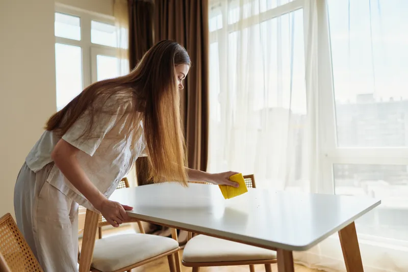 Woman cleaning table with yellow sponge in sunlit dining room, wiping surface during morning tidy up. Home interior with wooden chair, casual clothing and long hair braid.