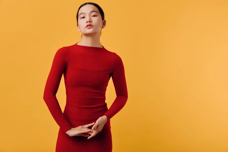 Young woman posing in red dress with long sleeves standing against orange background. Fashion model with calm expression and minimal makeup wearing fitted outfit for style photo shoot.