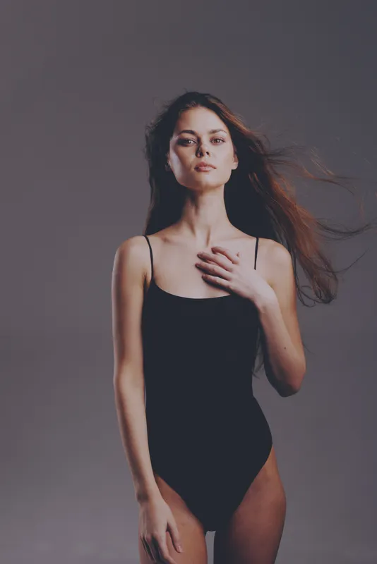 Young woman in black swimsuit posing confidently with wind-blown hair on gray background