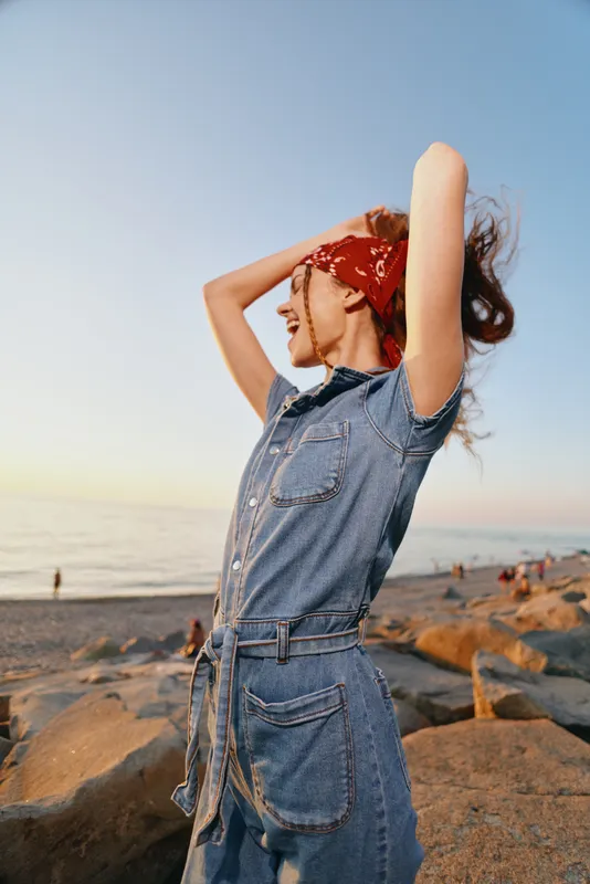 Lifestyle woman in modern boho-western denim street style stands on rocky beach at sunset, wearing red bandana and film color tones, enjoying warm evening breeze.