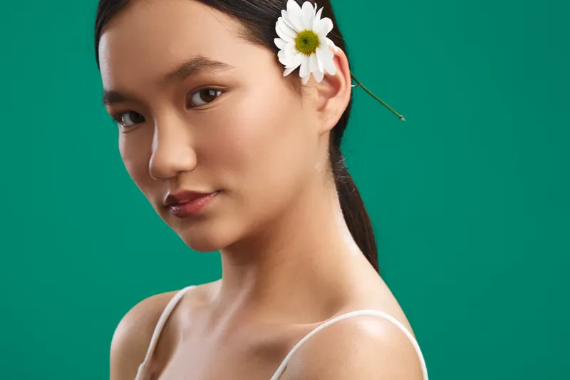 asian woman with a flower in her hair stands against a vibrant green backdrop, presenting a calm and elegant portrait that highlights natural beauty and poise.