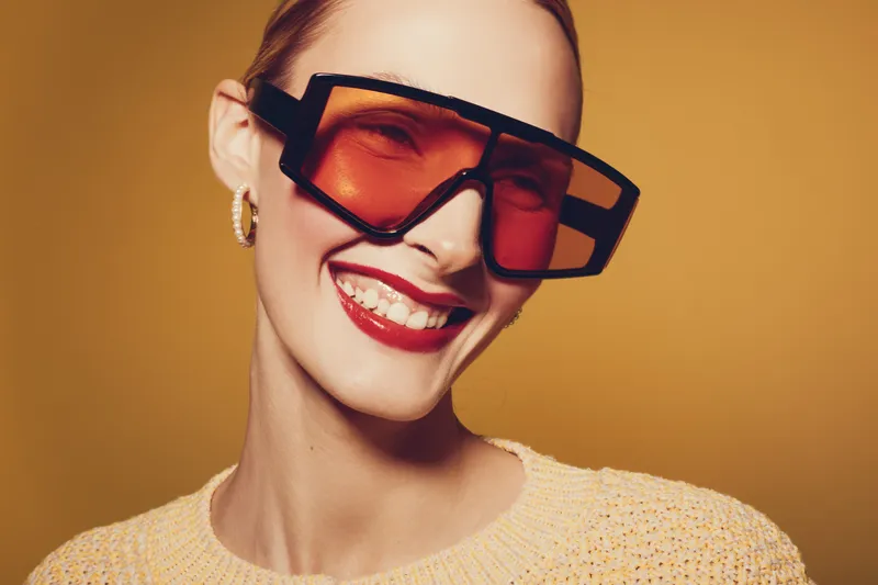 Smiling woman with red lipstick wearing oversized orange sunglasses and yellow sweater poses against warm brown background. Concept of fashion, style, confidence, and modern accessories.