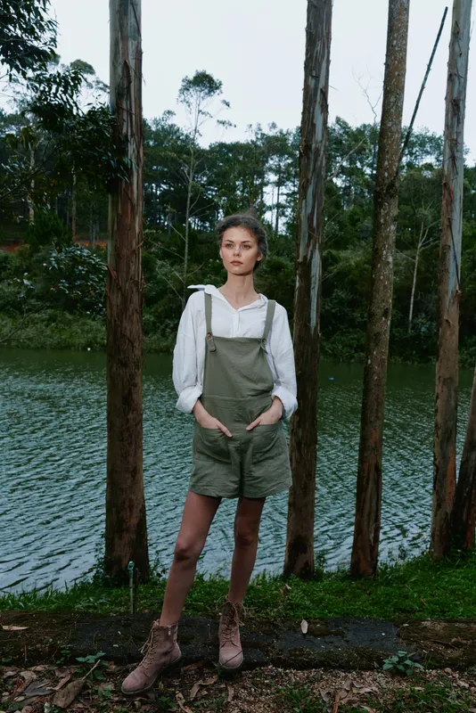 A young woman in casual green overalls stands by a serene lake, showcasing a natural vibe and peaceful expression amidst lush greenery and tall trees