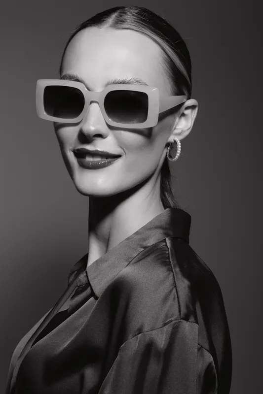 Stylish Caucasian woman with fair skin wears large square sunglasses and hoop earrings. She poses confidently in a dark silky blouse against grey background. Fashion concept.