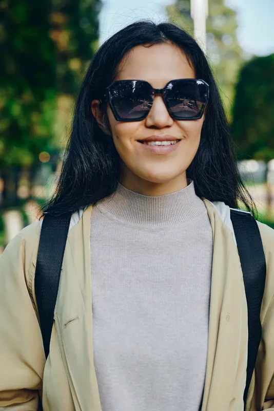Woman wearing sunglasses and beige coat outdoors, smiling in a sunny park, carrying a backpack for casual city style and travel mood