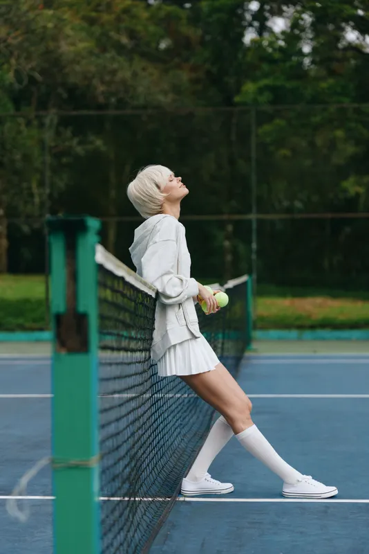Stylish young female tennis player enjoying a moment on the court, wearing a light athletic outfit, white skirt, and knee high socks, surrounded by greenery