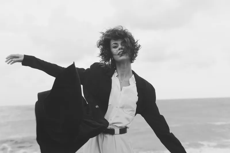 Fashion, wind, and beach scene with a flowing coat and crisp shirt, monochrome tones, dynamic pose, and open space, illustrating movement, style, and freedom in bold outdoor fashion photography