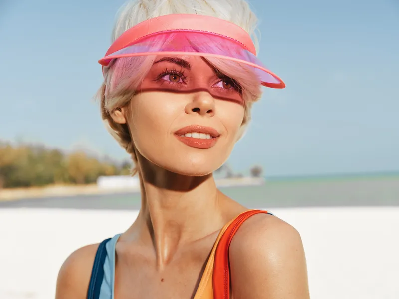 A stylish woman at the beach wears a translucent pink visor, colorful swimsuit, and radiant makeup, bathed in bright sunlight for a vivid summer portrait.