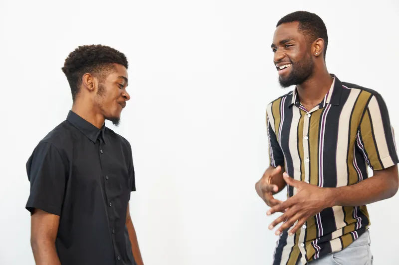 Two black men engaged in a lively conversation, expressing friendship and joy. The vibrant atmosphere is enhanced by their stylish clothing and warm smiles.