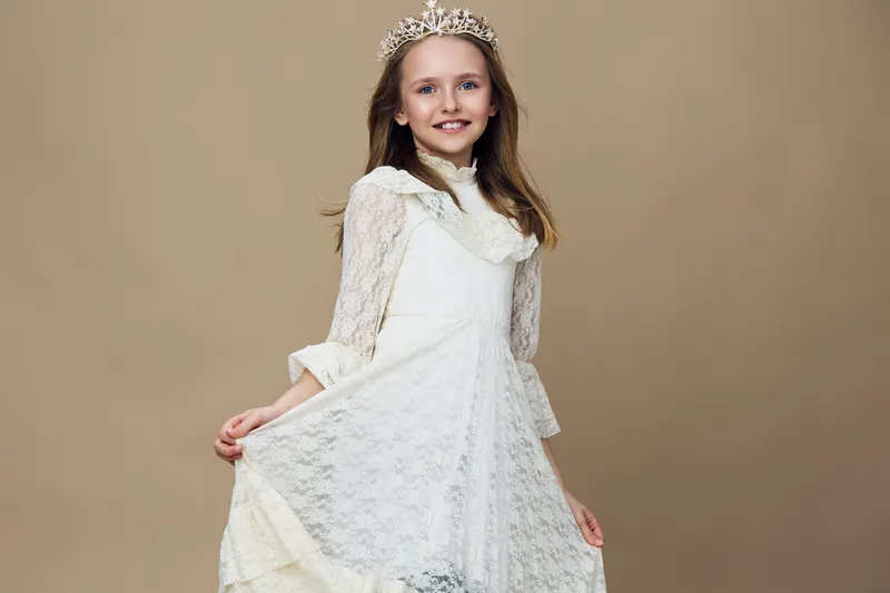 Young girl in elegant white dress with lace details and tiara, smiling and posing against a soft beige background, embodying innocence and joy
