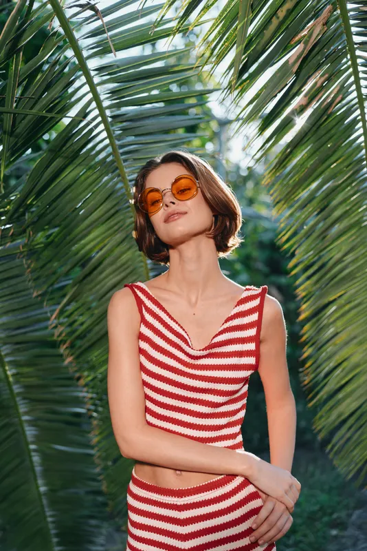 A cheerful woman poses in a tropical setting, wearing a red and white striped dress and round orange sunglasses. She exudes confidence and relaxation among lush palm leaves.