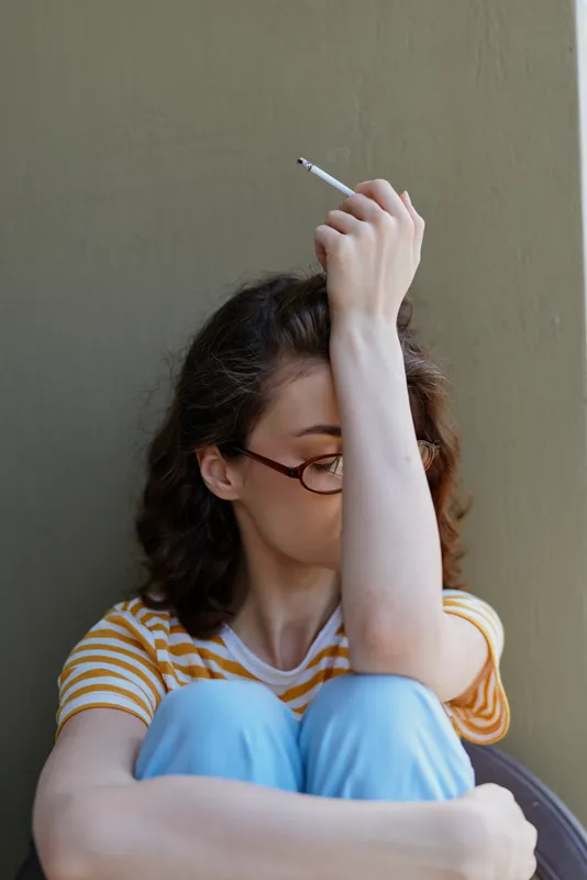 Reflective woman with striped shirt and glasses sits against a plain wall, one arm raised holding a cigarette, casual home setting, moody intimate moment, personal contemplation and quiet solitude.
