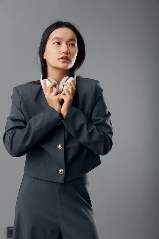 Young woman in business suit holding headphones with hands looking aside on grey background. Professional female model posing with casual headphones for work or leisure indoors.