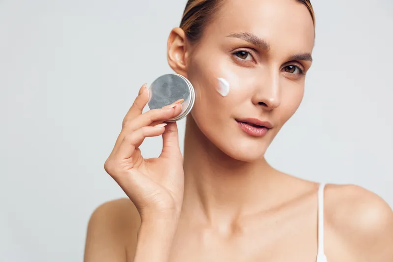 Young woman applies moisturizing cream on face holds jar of skincare product looks at camera with natural makeup isolated on light background closeup portrait beauty concept