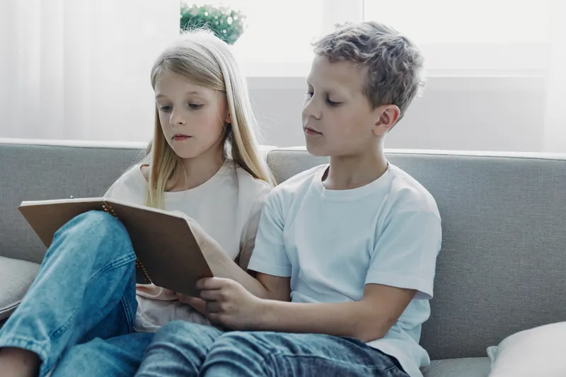 Two children sitting together on a sofa reading books in a bright living room. The boy and girl focus on their reading material in a calm, educational, and cozy home environment.