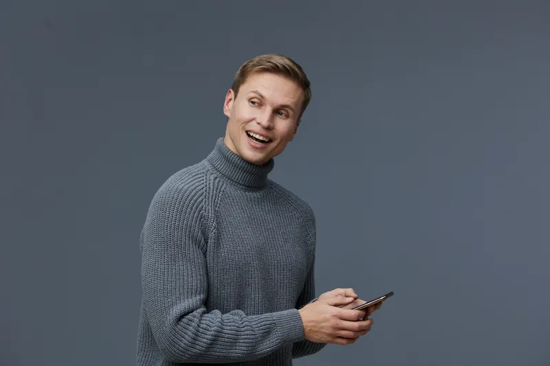 Smiling man using mobile phone, casual gray sweater, cheerful expression, trendy hairstyle, modern appearance, isolated on gray background, studio portrait, lifestyle concept.