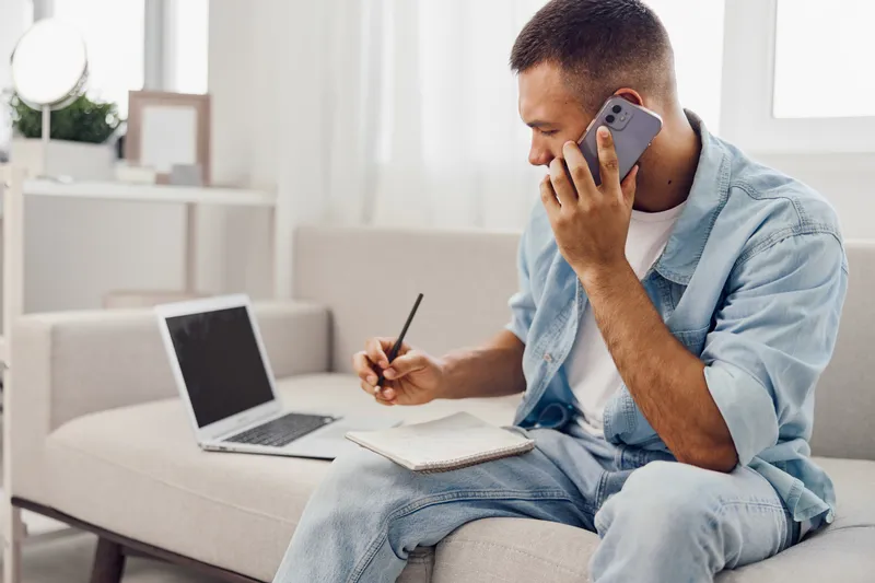 Casual man engaged in a phone call while jotting down notes, showcasing a modern workspace. Ideal for themes of productivity and remote work in 2025-2026.