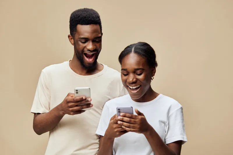 Smiling young African couple enjoying their smartphones together in a warm toned setting, showcasing joy and connection through technology