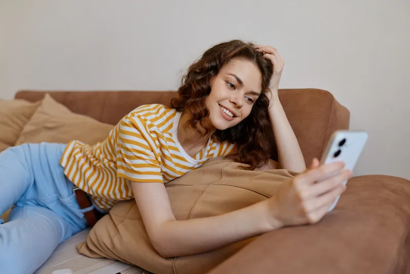 Cheerful woman indoors, lounging on a sofa, smiling at phone, relaxed lifestyle moment, soft natural light, cozy home setting, casual fashion, warm colors, modern living