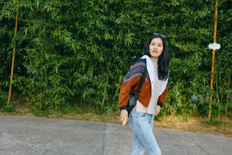 Woman walking along a street in casual fashion, wearing a patterned cardigan and jeans, with lush greenery in the background, showcasing street style and relaxed confidence outdoors