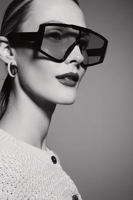 Stylish white woman with pale skin wearing large sunglasses and earrings poses in studio with knitted top. Fashion portrait with minimalistic lighting.