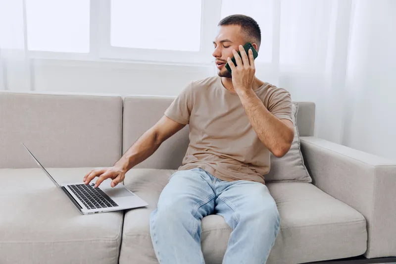 Smiling man engaged in a phone conversation while working on a laptop, showcasing modern remote work culture and a relaxed home environment.