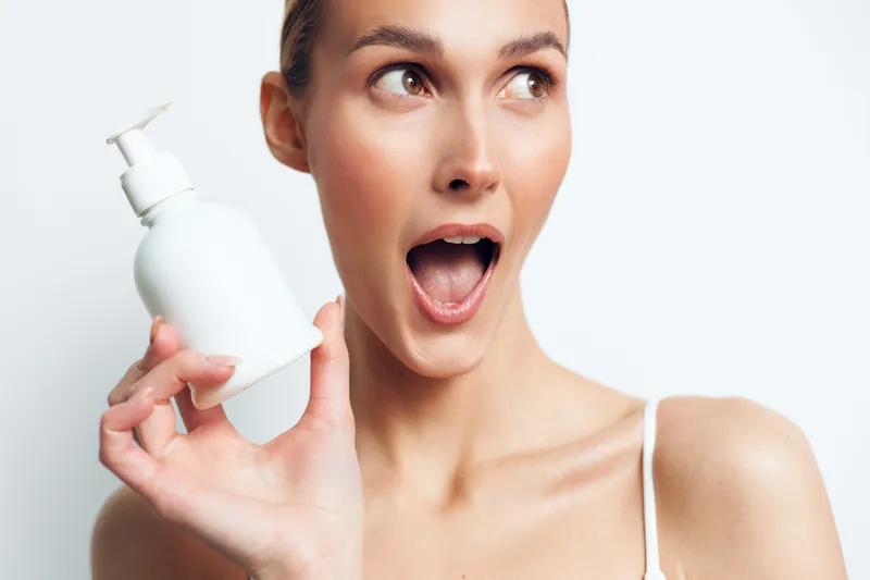 A surprised young woman with clear skin holds a white cosmetic bottle near her face against a plain white background. Concept for skincare, beauty, and personal care products.